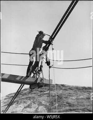 A rigger attaching suspenders to a pair of the 2-1/2 inch suspension cables of the sand and gravel conveyor bridge.; Scope and content:  Photograph from Volume Two of a series of photo albums documenting the construction of the Grand Coulee Dam and related work on the Columbia Basin Project. Stock Photo