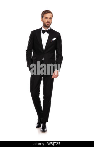 relaxed businessman in black suit looks to side while walking on white background with a hand in pocket Stock Photo