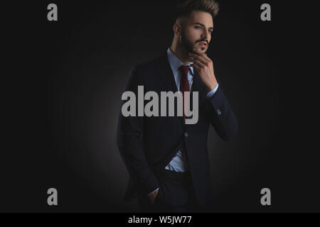 portrait of pensive and relaxed businessman in navy suit and red tie looking down to side while standing on black background Stock Photo