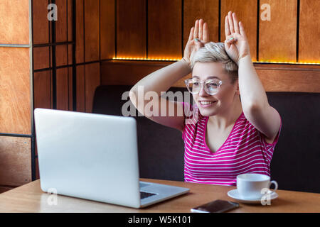Happy childish young girl with blonde short hair in pink shirt is sitting in cafe and making video call on laptop, talking and teased with her friend, Stock Photo