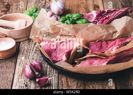 Raw veal steaks with cooking  ingredients, sea salt, garlic, basil herbs ready to cook, preparing food, rural wooden table, close up, selective focus Stock Photo