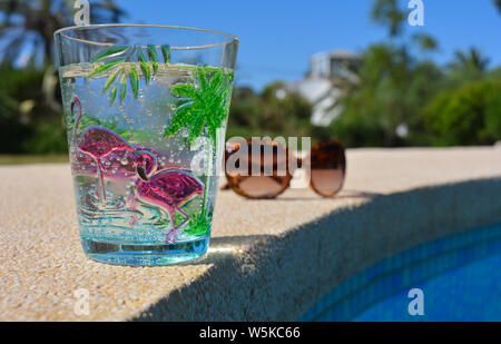 Pool day, summer fun vibes. Sparkling cold water in a tropical themed plastic cup and plastic tortoiseshell  coloured sunglasses on the poolside Stock Photo