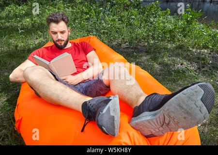 Young bearded student is reading a book lying on the orange air sofa on grass in forest during eco-tourism. Stock Photo