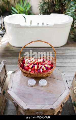 Wooden wicker basket with red and white flower petals in front of white stone empty bath over green tropical nature background. Spa relaxation concept Stock Photo