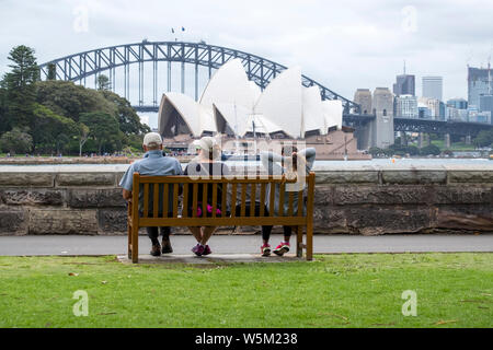 Three people sit on a bench looking out to the Sydney Opera House and Sydney Harbour Bridge from Farm Cove, Sydney Stock Photo