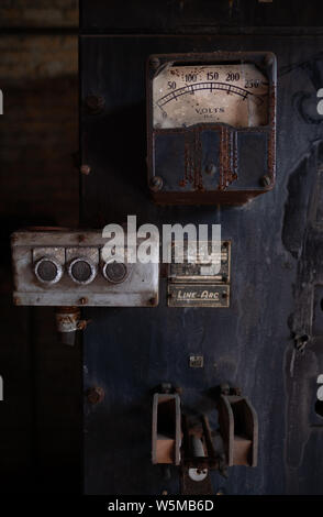 Vintage industrial electrical control panel with rusted analog voltmeter gauge and mechanical switches in an abandoned factory Stock Photo