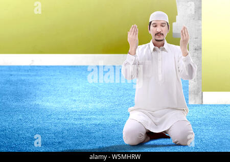 Asian Muslim man sitting in pray position while raised hands and praying inside the mosque Stock Photo