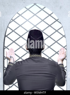 Rear view of Asian Muslim man standing and praying with prayer beads while raised arms inside the mosque Stock Photo