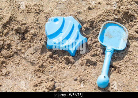 Blue sand toys, a shovel and a mold of a fish, are lying in the sand on a sunny beach in the summertime, waiting to be played with. Stock Photo