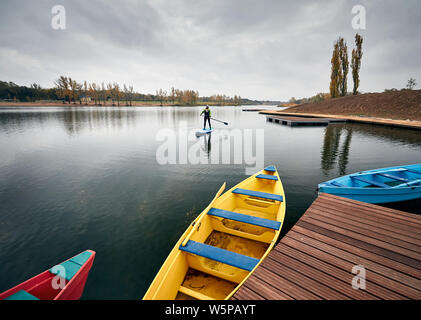 Man on the paddleboard in the lake against overcast sky and pier with colorful boats at foreground Stock Photo