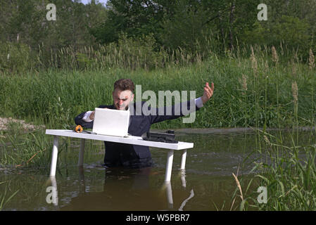 Young bearded millennial man, guy tired of office work. Angry and distracted in the workplace. At the table, standing in the swamp, in the water Stock Photo