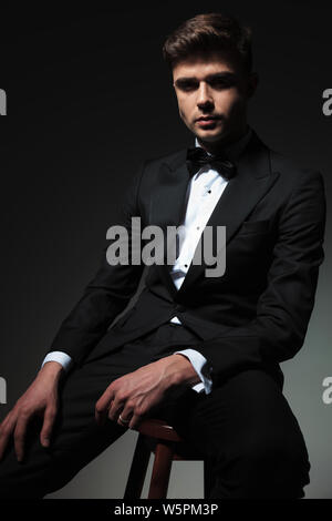 elegant young man in tuxedo sits on wooden chair on grey background while resting hand on thigh Stock Photo