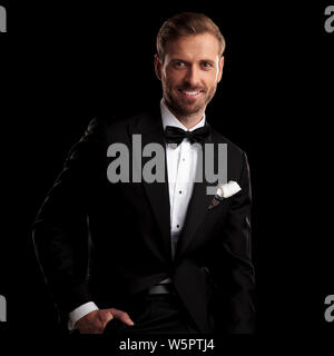 smiling young man in tuxedo with hand in pocket is sitting against black background Stock Photo