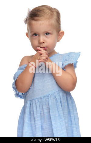 cute little girl prays with palms together and one finger in her mouth on  white background Stock Photo