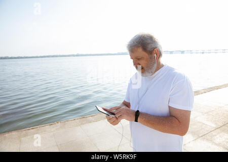Senior man as runner with fitness tracker at the riverside. Caucasian male model using gadgets while jogging and cardio training in summer's morning. Healthy lifestyle, sport, activity concept. Stock Photo
