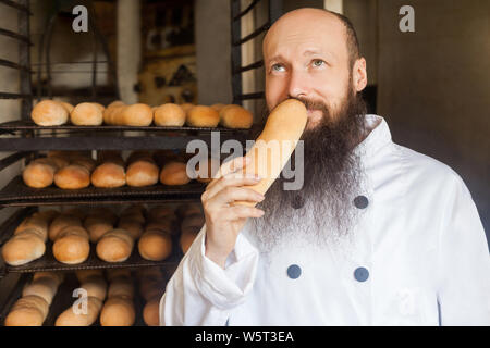 Portrait of cheerful young adult baker with long beard in white uniform standing in his manufacture, smelling with pleasure fresh bread and enjoying. Stock Photo