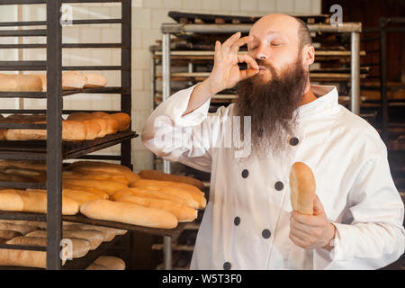Portrait of professional adult male chef with long beard in white uniform standing in his workplace and cook with taste approval gesture, showing perf Stock Photo