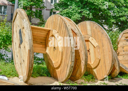 Large broken empty coils and reels with wooden cable drums, stacked on an open street area with sand and grass in the city park near to the houses Stock Photo