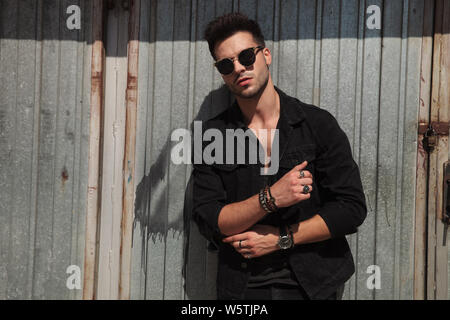 portrait of casual man with sunglasses fixing his black shirt's sleeves while standing near metal door outside Stock Photo