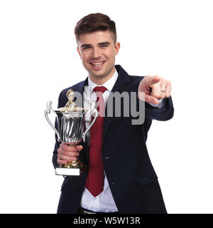 portrait of smiling businessman pointing finger while holding the awarded trophy. He is standing on white background while wearing a red tie and a nav Stock Photo