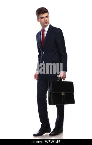 businessman with briefcase in hand looks to side while walking on a white background. He wears a navy suit and a red tie, full body picture. Stock Photo