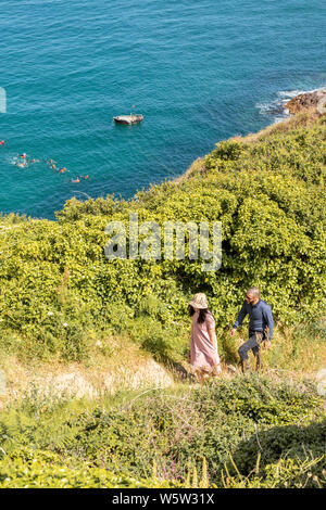 A young couple walk the coastal path while a group of young people are coasteering below the cliffs at Pointe de la Moye, Le Gouffre, Les Villets on t Stock Photo