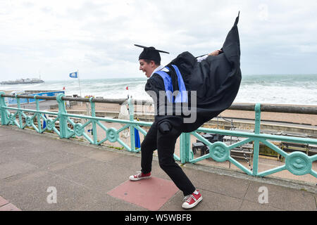 Brighton, UK. 30th July, 2019. Emily Green hangs on to her gown after graduating from the University of Brighton as she celebrates on a very windy seafront afterwards . Credit: Simon Dack/Alamy Live News Stock Photo