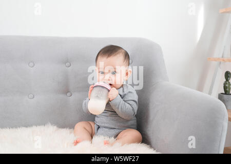 Little cute baby girl sitting in room on sofa drinking milk from bottle and smiling. Happy infant. Family people indoor Interior concepts. Childhood b Stock Photo