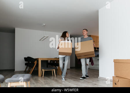 Couple moving into new house. Cheerful young man and woman carrying cardboard boxes in half empty living room. Stock Photo