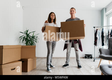 Couple moving into apartment together. Cheerful young man and woman looking at camera holding cardboard boxes in room. Stock Photo