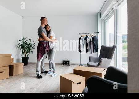 Happy couple hugging together in new house. Moving in together - young cheerful man and woman cuddling in room with cardboard boxes. Stock Photo