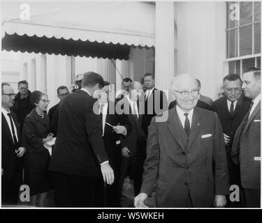 Photograph of former President Harry S. Truman smiling during his visit to the White House, as President John F. Kennedy confers with an aide in the background. Stock Photo