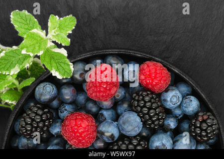 Composition of pineapple mint green twigs, blueberries, blackberries and raspberries in round and black cast iron bowl on black stone background with Stock Photo