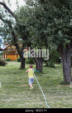 Boy pulling a garden hose to watering the plants in backyard garden. Candid people, real moments, authentic situations Stock Photo