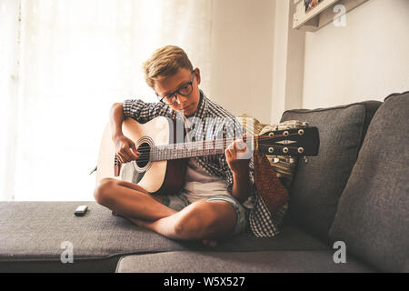 Beautiful young boy playing acoustic guitar. Music student exercises alone at home. Trendy caucasian child plays instruments sitting legs crossed on t Stock Photo