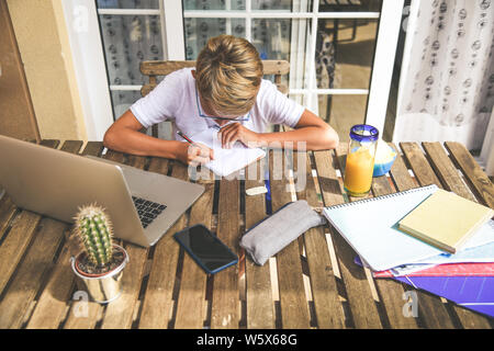 Beautiful young boy doing homework outdoor. Child working with pencil and laptop at home after school. Kid studying on the terrace during holiday drin Stock Photo