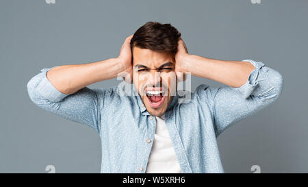 Emotional Hispanic Man Screaming Covering His Ears On Gray Background Stock Photo