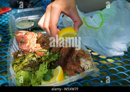 Hand squeezing a freshly cut lemon over a juicy take out fried red snapper. Stock Photo