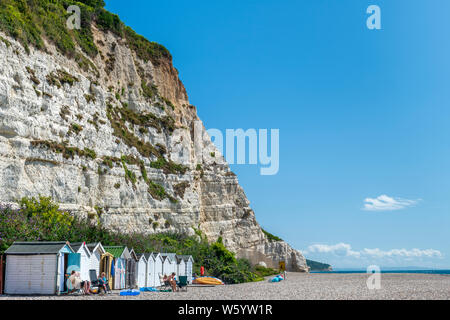 Beer, South East Devon, England. Holidaymakers enjoy the summer sunshine beside the little huts that sit beneath the cliffs on the beach at Beer in So Stock Photo