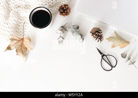 Autumn, fall workspace composition with closed laptop. Cup of coffee, wool blanket, autumn leaves, dry eucalyptus, scissors and pine cones on white ta Stock Photo