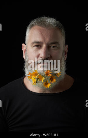Portrait of smiling brutal man with a gray beard decorated with yellow flowers on a black background, selective focus Stock Photo