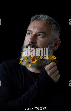 Portrait of pensive brutal man with a gray beard decorated with yellow flowers on a black background, selective focus Stock Photo