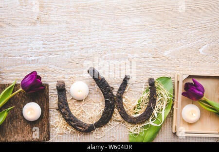 Flat lay view of two rusty small and big horseshoes on light wooden board background, decorated with white small candles and purple tulips, spring in Stock Photo