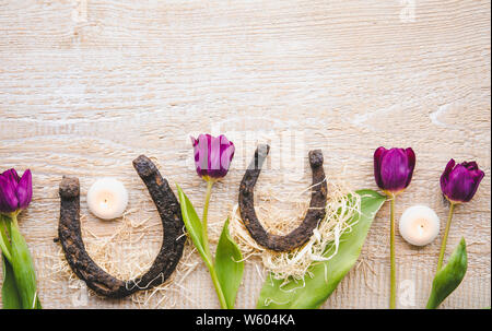 Flat lay view of two rusty small and big horseshoes on light wooden board background, decorated with white small candles and purple tulips, spring in Stock Photo