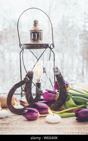 Old rusty horseshoes, brown vintage lantern with modern dimmable decorative light bulb switched on and white candles lit and  tulips on wooden window Stock Photo