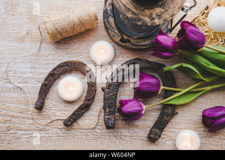 Flat lay view of two rusty small and big horseshoes on light wooden board background, decorated with white small candles and purple tulips, spring in Stock Photo