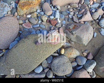 Various shapes and sizes of rocks, stones and pebbles in colors of coral, jade, rust, and tan in bright sunlight scattered on a dark brown sandy beach Stock Photo