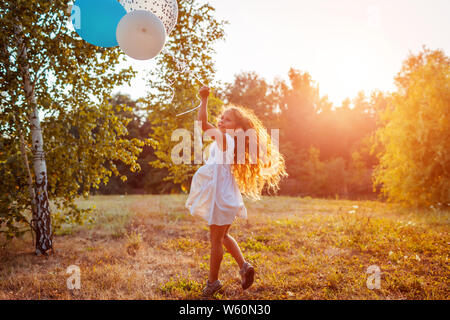 Happy little girl running with baloons in hand. Kid having fun in summer park at sunset. Stock Photo