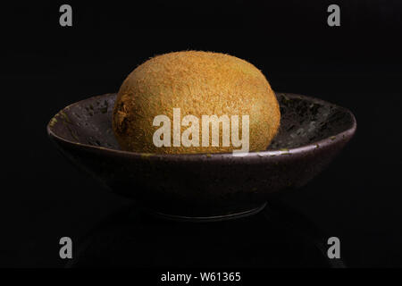 One whole fresh green kiwifruit actinidia deliciosa in a dark ceramic bowl isolated on black glass Stock Photo