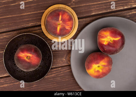 Group of four whole fresh red peach on a round bamboo coaster in a dark ceramic bowl on a gray ceramic plate flatlay on brown wood Stock Photo